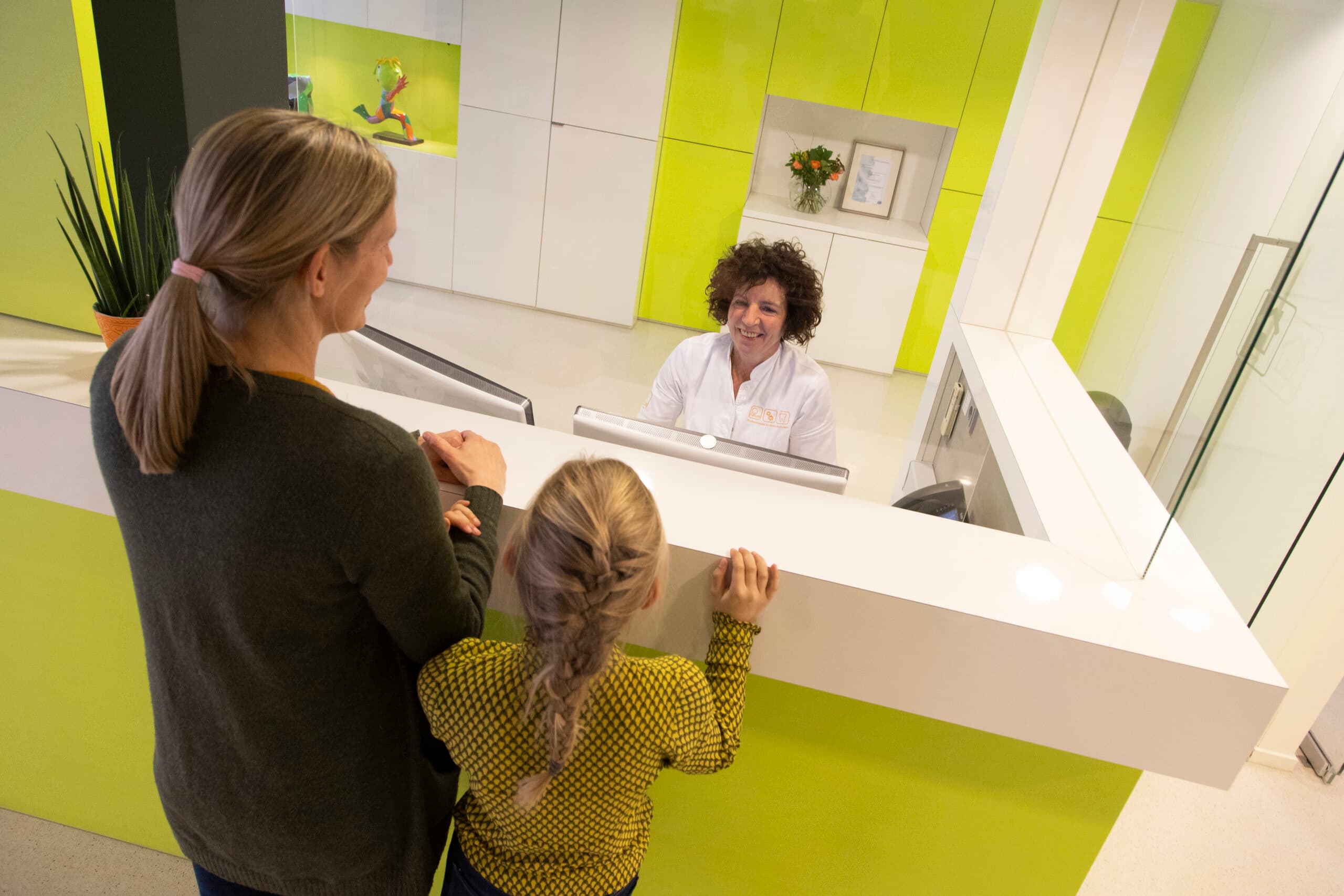 Een patiënt staat bij de balie van een orthodontist met twee kinderen. Achter de balie staat een vrouw die hen helpt.